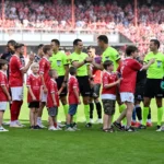 Voetballers en scheidsrechters staan opgesteld op het veld voor een wedstrijd.