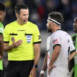 Een scheidsrechter spreekt op het veld met spelers tijdens een Belgische competitiewedstrijd.