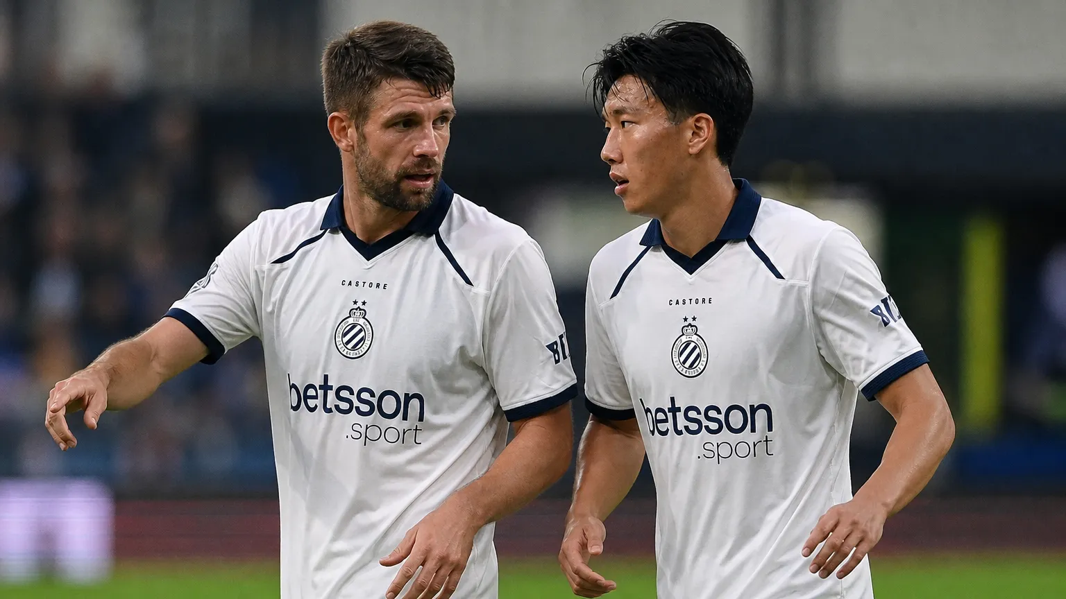 Twee Club Brugge-spelers in witte tenues overleggen tijdens een wedstrijdmoment.