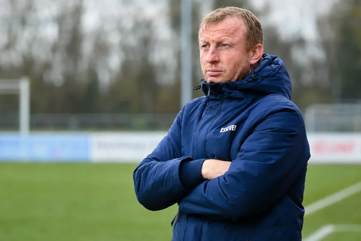 Sven Vandenbroeck in een blauwe coachjas aan de rand van een voetbalveld.