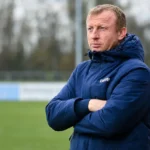 Sven Vandenbroeck in een blauwe coachjas aan de rand van een voetbalveld.