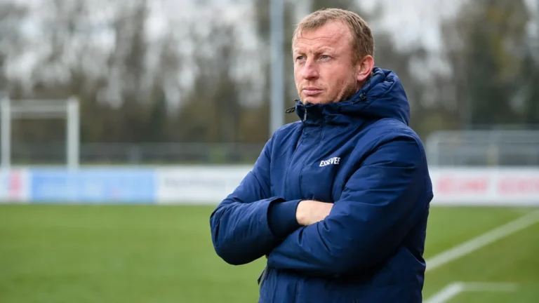 Sven Vandenbroeck in een blauwe coachjas aan de rand van een voetbalveld.