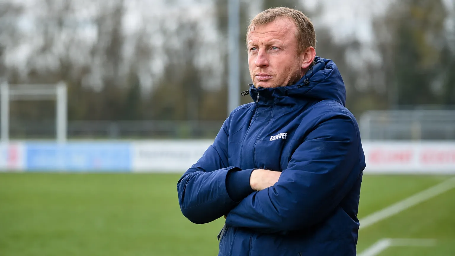 Sven Vandenbroeck in een blauwe coachjas aan de rand van een voetbalveld.