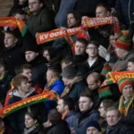 Collectieve supporters op een voetbaltribune met rood-geel-groene sjaals.