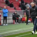 Een niet-spelende man, een jonge supporter in rode kleding en een fotograaf langs de zijlijn van een stadion.