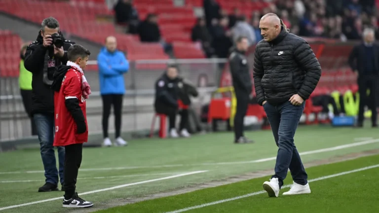 Een niet-spelende man, een jonge supporter in rode kleding en een fotograaf langs de zijlijn van een stadion.