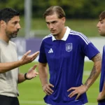 Een trainer geeft op het oefenveld aanwijzingen aan voetballers in paarse shirts.