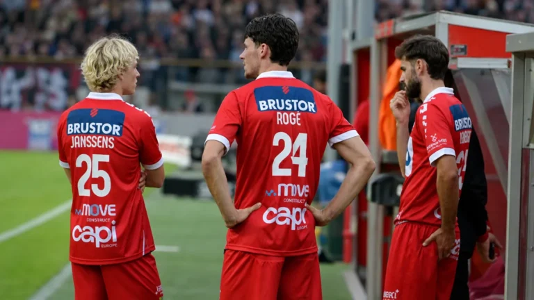 Een groep spelers in rode tenues wacht samen aan de zijlijn van een voetbalstadion.