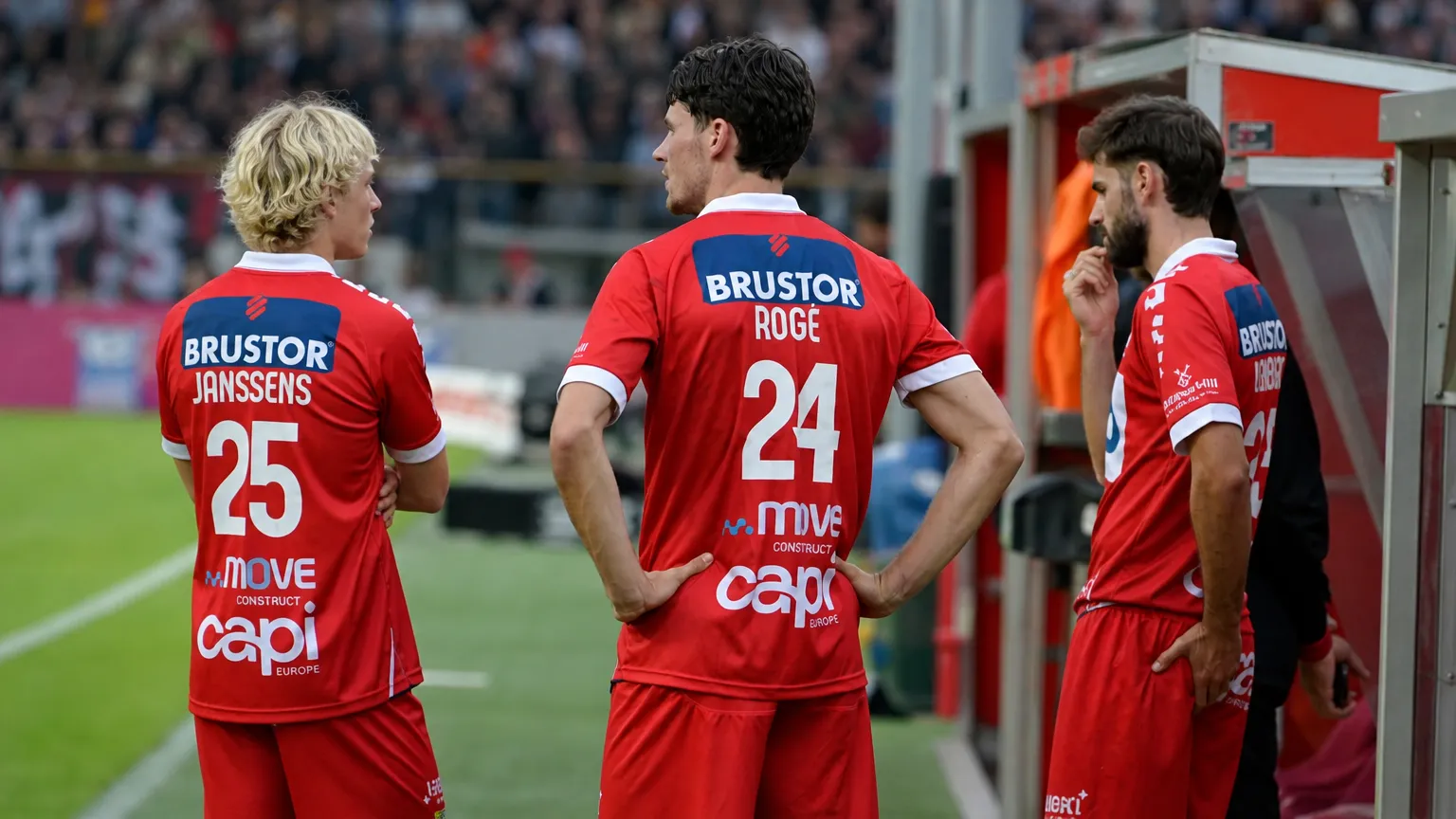 Een groep spelers in rode tenues wacht samen aan de zijlijn van een voetbalstadion.