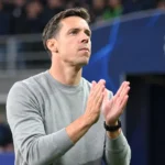Een coach in grijze trui applaudisseert langs de zijlijn van een voetbalstadion.