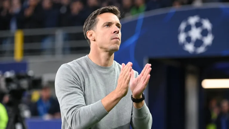 Een coach in grijze trui applaudisseert langs de zijlijn van een voetbalstadion.