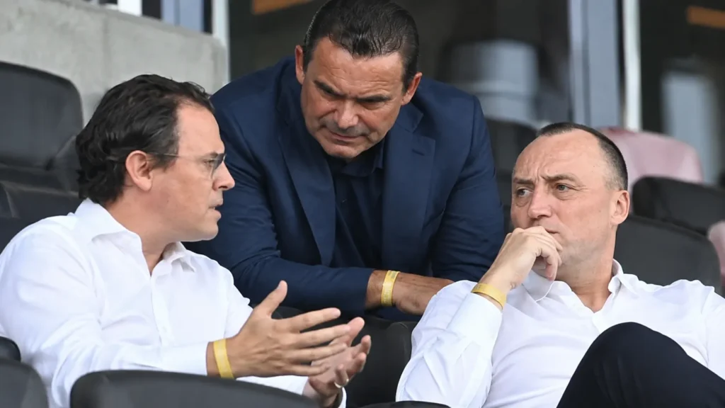 antwerp-peter-catteeuw-grotere-sportieve-rol-overmars Drie clubvertegenwoordigers in formele kledij op de zitplaatsen van een stadiontribune.
