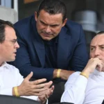 Drie clubvertegenwoordigers in formele kledij op de zitplaatsen van een stadiontribune.