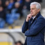 Een oudere voetbalcoach in een donkerblauwe jas kijkt geconcentreerd vanaf de zijlijn naar het veld.