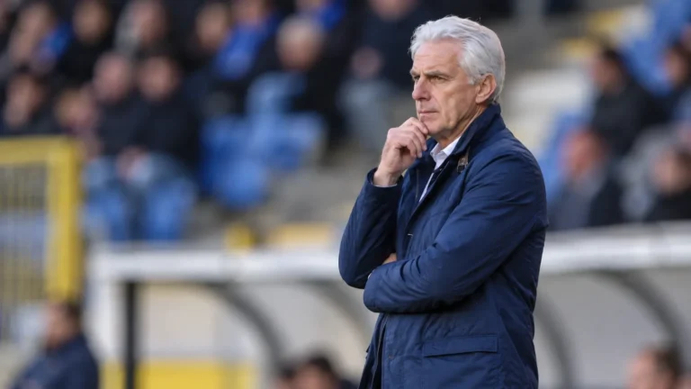 Een oudere voetbalcoach in een donkerblauwe jas kijkt geconcentreerd vanaf de zijlijn naar het veld.