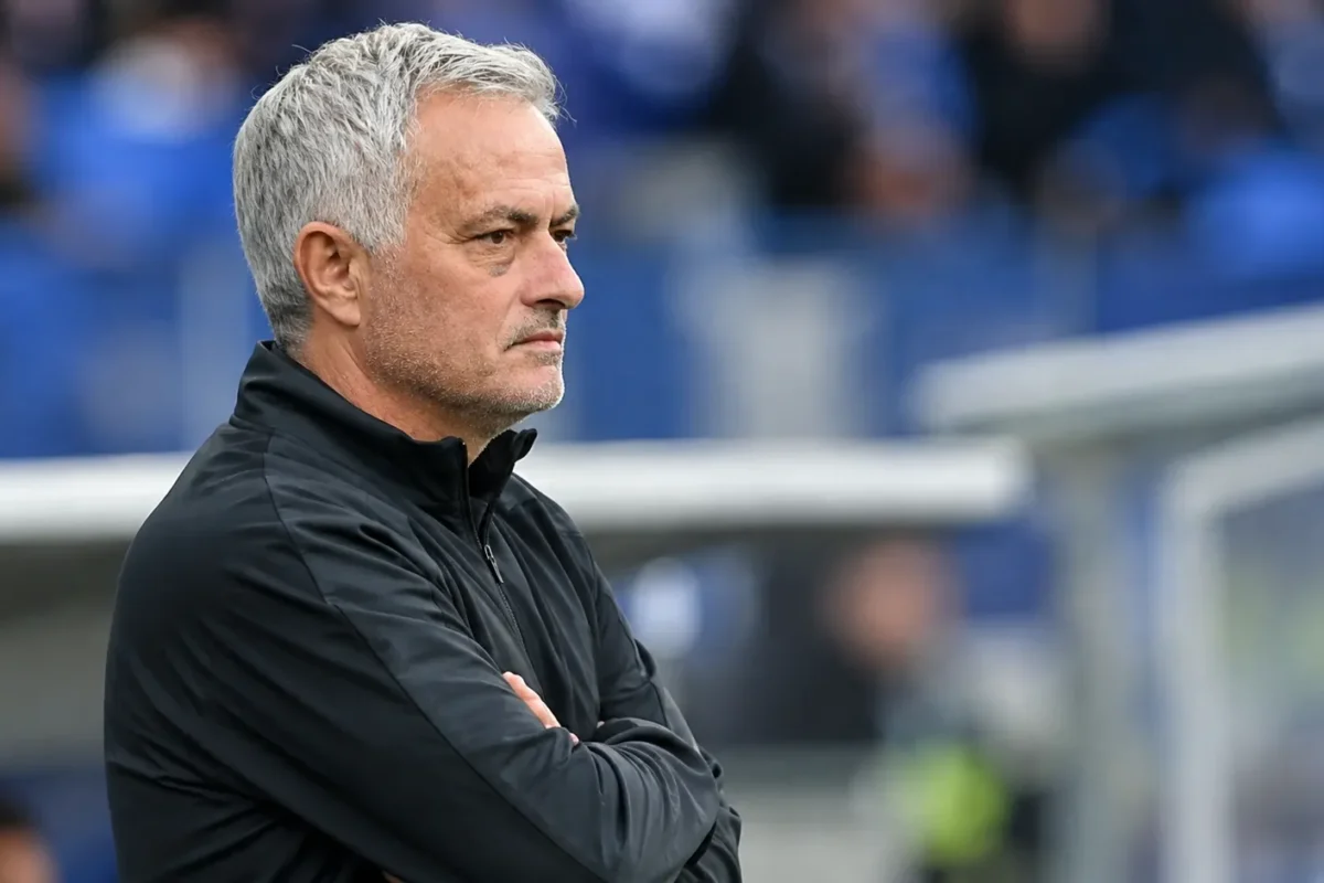 Een grijsgekleurde voetbalcoach kijkt geconcentreerd vanaf de zijlijn van een stadion.