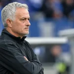 Een grijsgekleurde voetbalcoach kijkt geconcentreerd vanaf de zijlijn van een stadion.