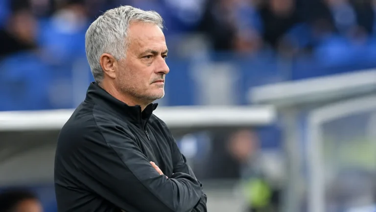 Een grijsgekleurde voetbalcoach kijkt geconcentreerd vanaf de zijlijn van een stadion.