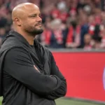 Een niet-spelende clubvertegenwoordiger in zwarte kleding langs de zijlijn, met een Bayern München-bord op de achtergrond.
