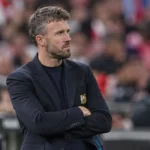 Een coach in donker jasje staat geconcentreerd langs de zijlijn van een voetbalstadion.