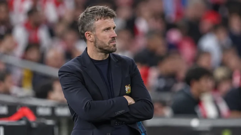 Een coach in donker jasje staat geconcentreerd langs de zijlijn van een voetbalstadion.
