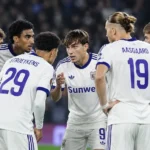 Een groep voetballers in witte tenues overlegt op het veld tijdens een avondwedstrijd.