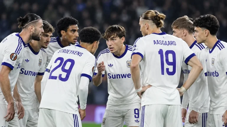 Een groep voetballers in witte tenues overlegt op het veld tijdens een avondwedstrijd.