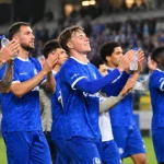 Een groep voetballers in blauwe wedstrijdshirts applaudisseert na afloop richting de tribunes.