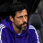 Een coach in paarse sporttrui zit geconcentreerd in een Europese wedstrijddug-out.