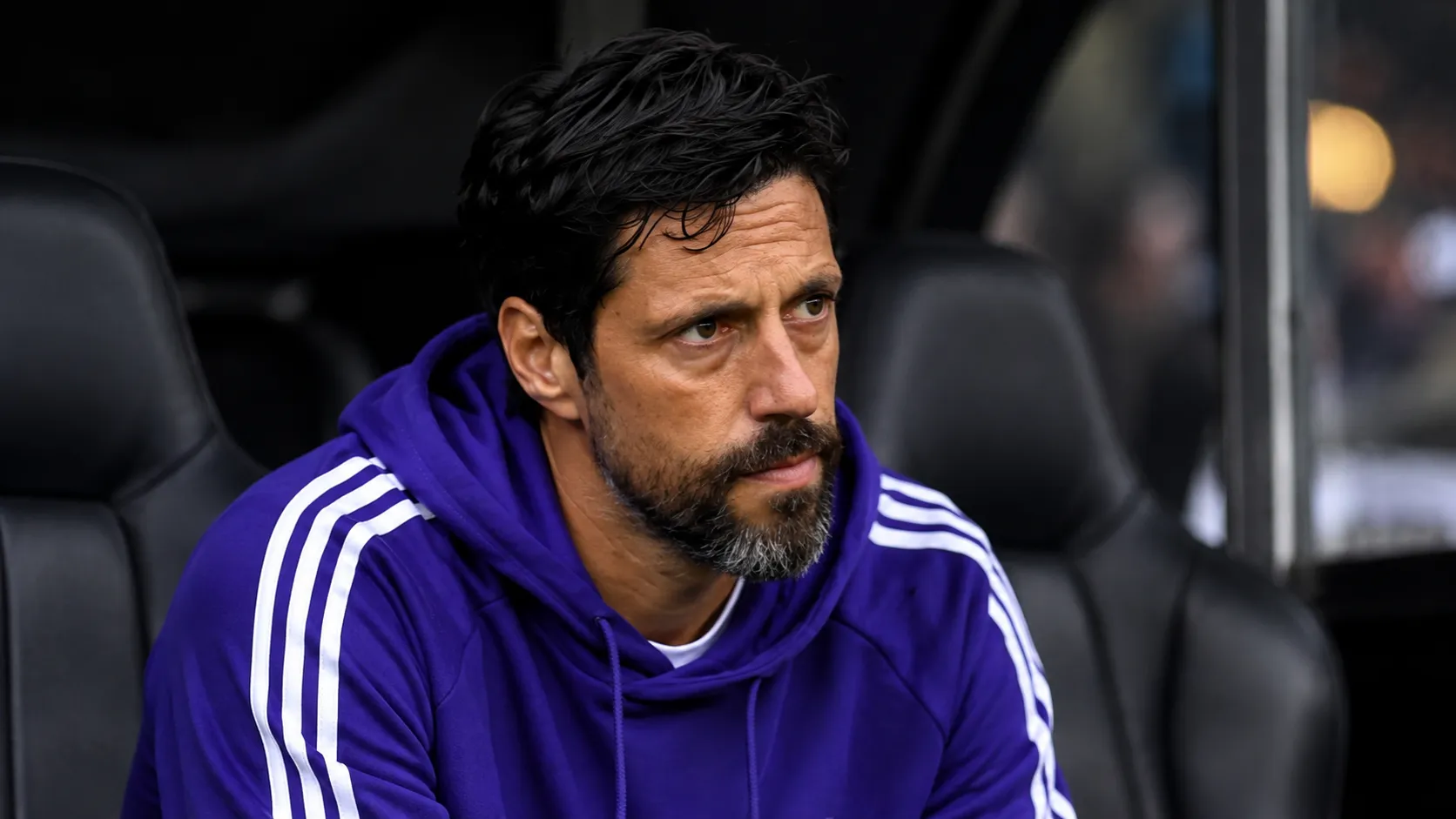Een coach in paarse sporttrui zit geconcentreerd in een Europese wedstrijddug-out.