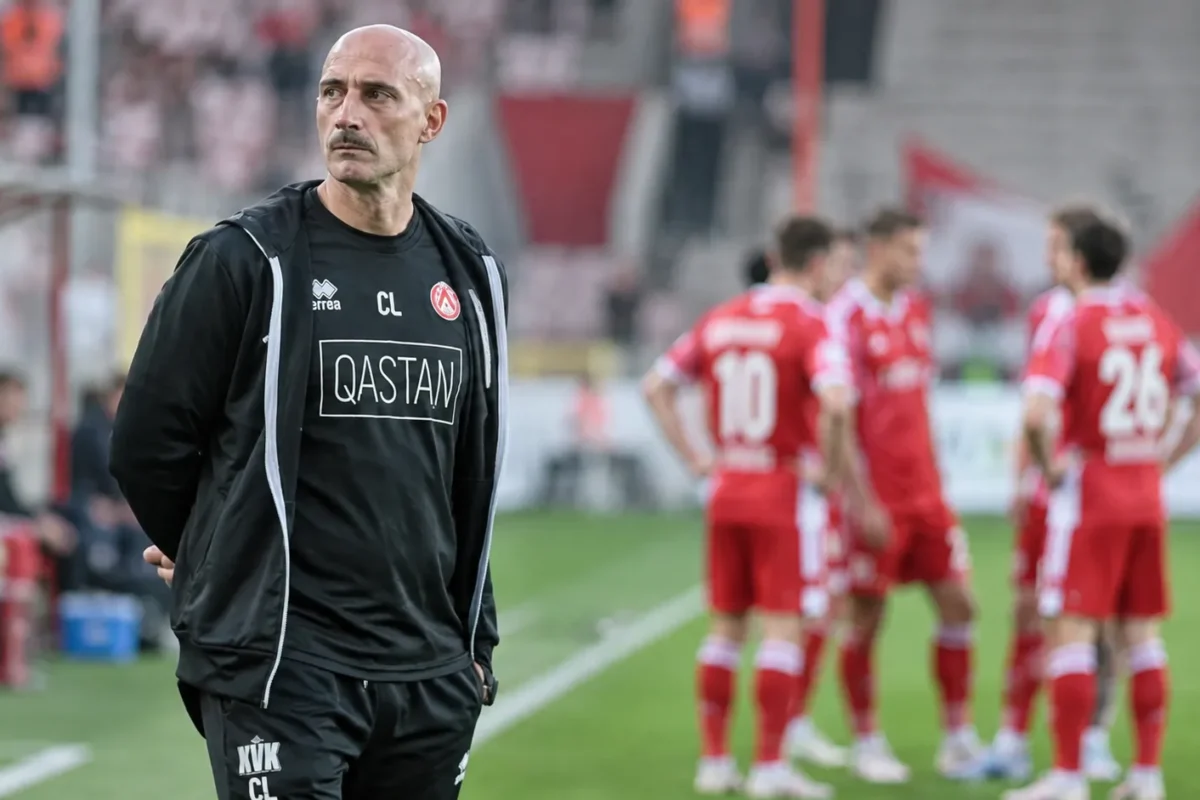 Een clubmedewerker in donkere sportkleding aan de zijlijn, met spelers op de achtergrond.