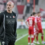 Een clubmedewerker in donkere sportkleding aan de zijlijn, met spelers op de achtergrond.