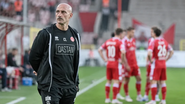 Een clubmedewerker in donkere sportkleding aan de zijlijn, met spelers op de achtergrond.