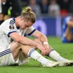 Een ontgoochelde voetballer in een wit tenue zit na de wedstrijd op het veld.