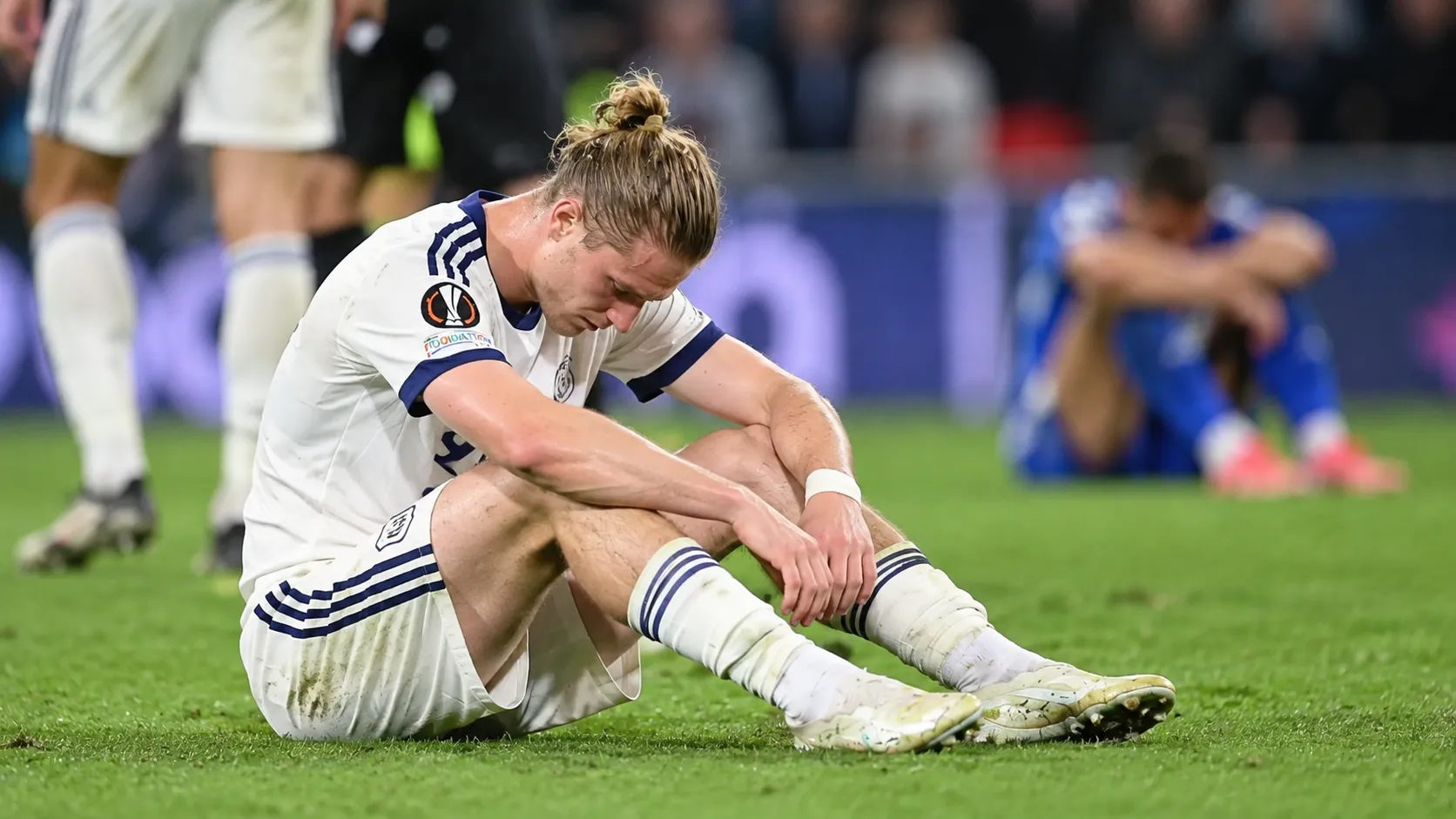 Een ontgoochelde voetballer in een wit tenue zit na de wedstrijd op het veld.