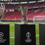 Drie Europese voetbaltrofeeën op zwarte sokkels langs een grasveld in een stadion.
