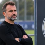 Een coach in een zwarte trui kijkt toe aan het trainingsveld van Club Brugge, met een clubembleem op een vast bord naast het veld.