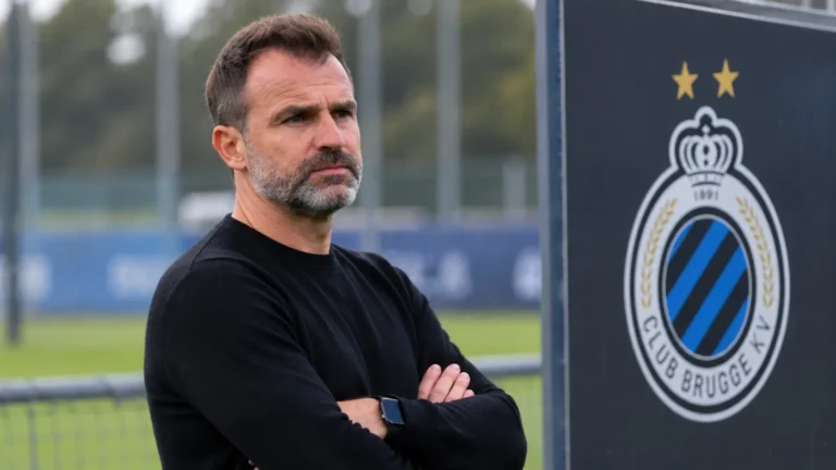 Een coach in een zwarte trui kijkt toe aan het trainingsveld van Club Brugge, met een clubembleem op een vast bord naast het veld.