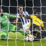 Spelers in geel en blauw-wit en een doelman in groen bij een verdedigende actie voor doel.