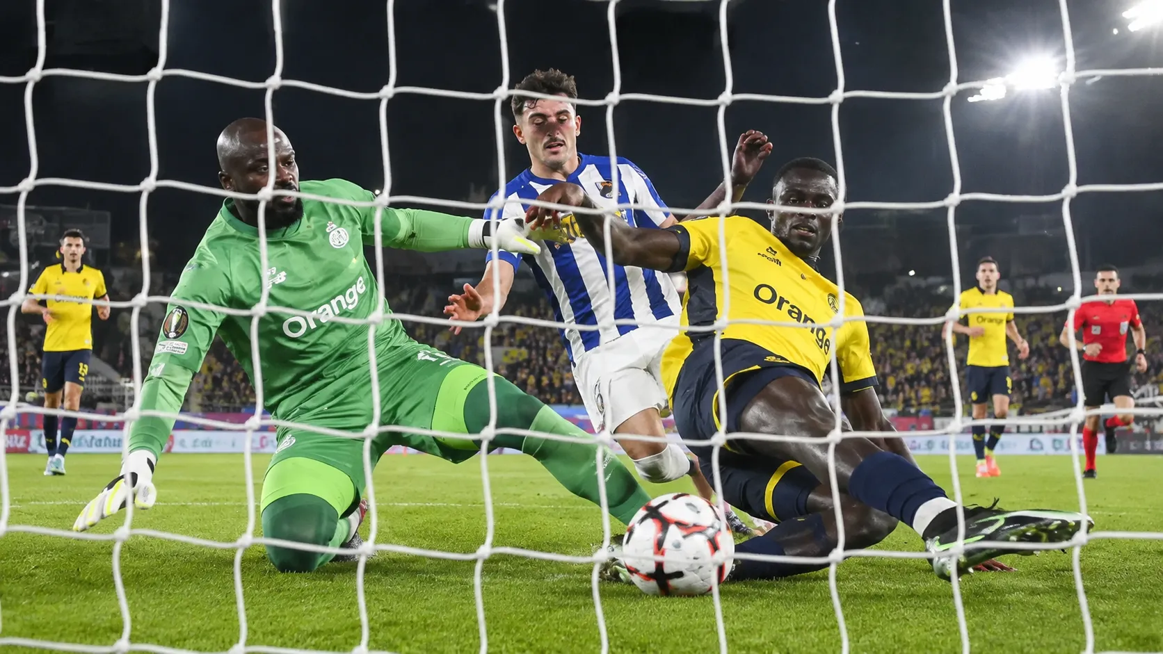 Spelers in geel en blauw-wit en een doelman in groen bij een verdedigende actie voor doel.