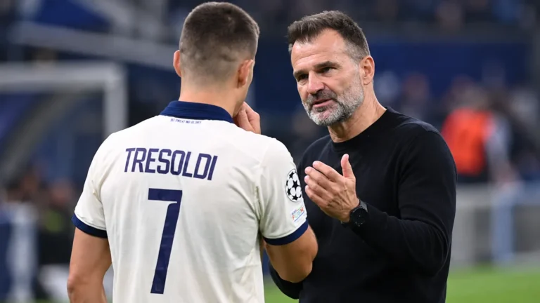 Een Club Brugge-trainer overlegt langs het veld met een speler in een licht wedstrijdshirt.