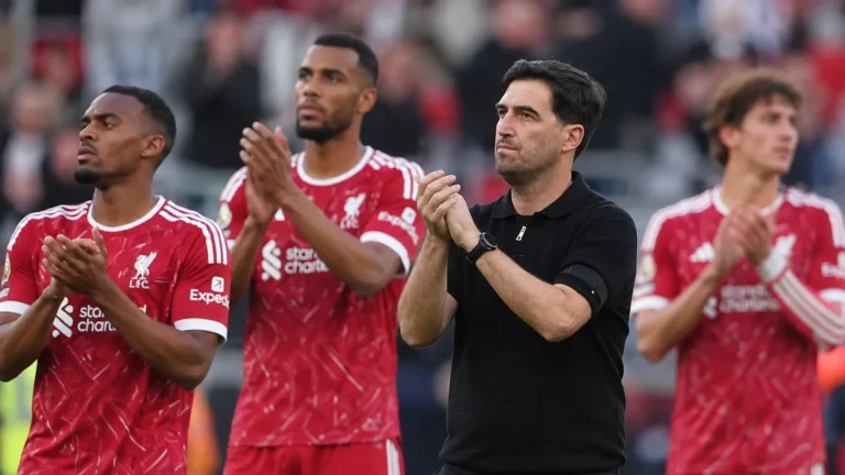 Een staflid en enkele spelers applaudisseren langs de zijlijn van een voetbalstadion.