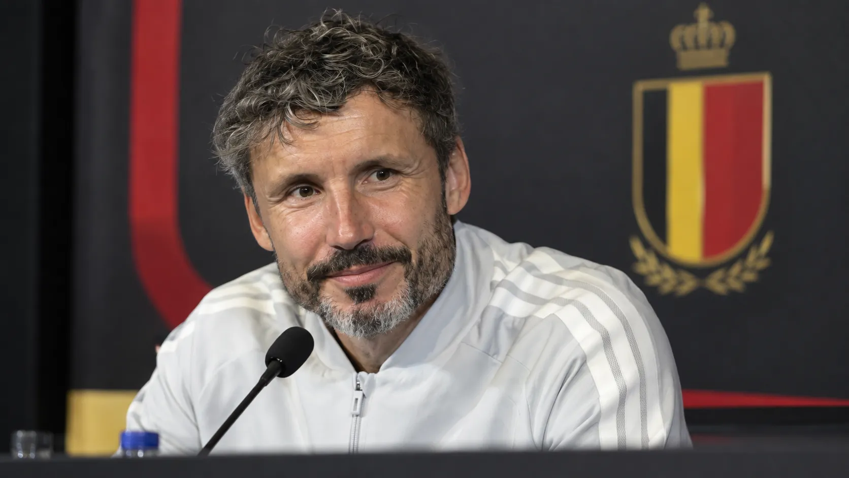 Mark van Bommel aan een persconferentietafel met een Belgisch nationaal ploegembleem op de achtergrond.