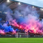 Een volle voetbaltribune met supporters tussen blauwe en roze rookwolken.