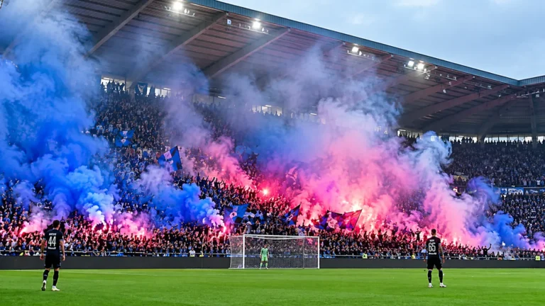 Een volle voetbaltribune met supporters tussen blauwe en roze rookwolken.