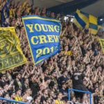 Een compacte groep supporters in gele en blauwe kledij zwaait met vlaggen op de tribune.