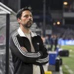 Een coach in donkere sportkleding naast de dug-out voor een bekerwedstrijd.
