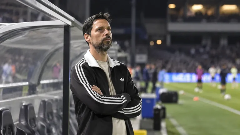 Een coach in donkere sportkleding naast de dug-out voor een bekerwedstrijd.