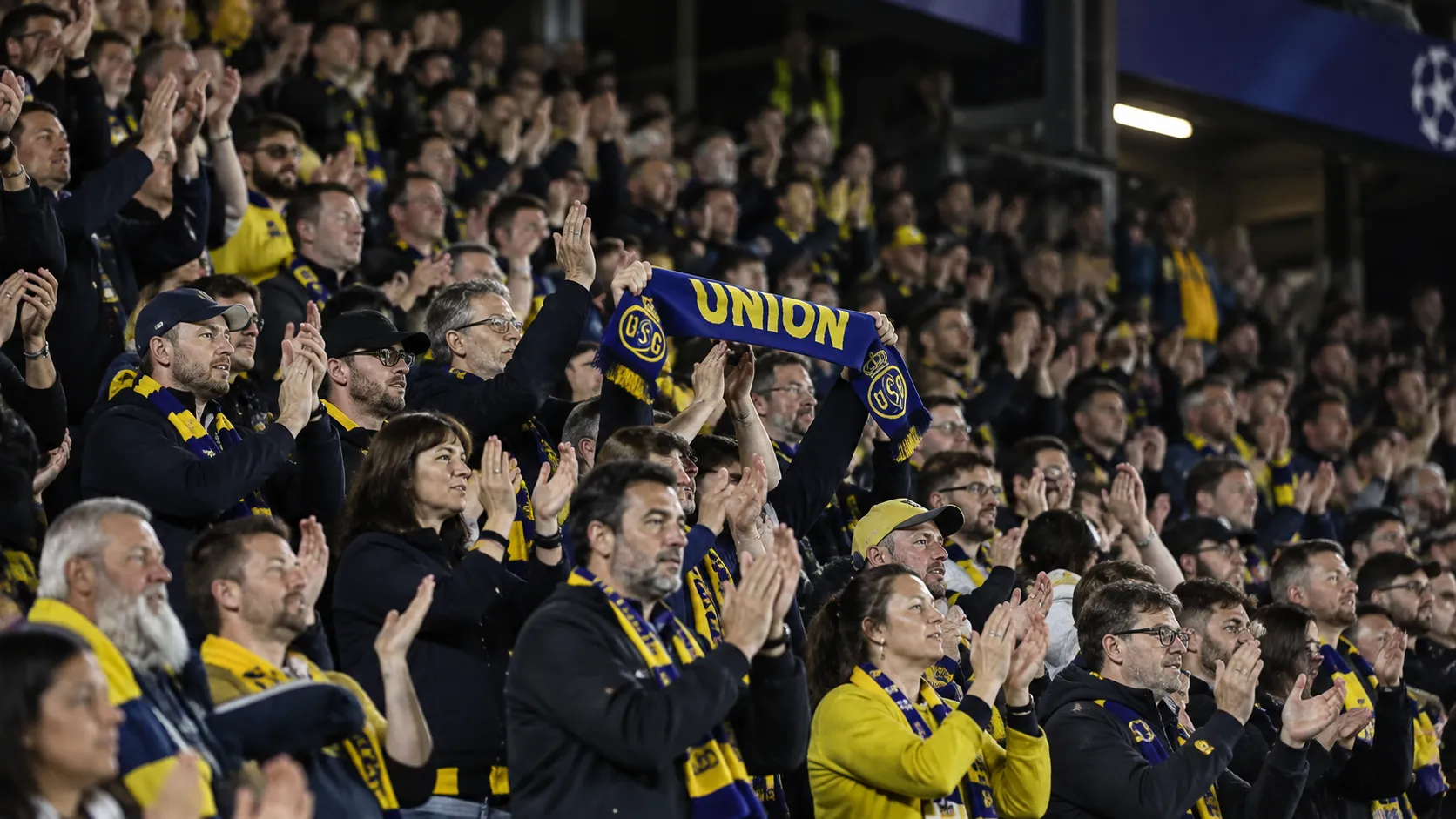 Supporters van Union applaudisseren in de tribune met een blauw-gele sjaal.