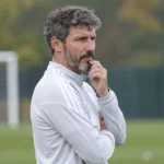 Een bondscoach in een witte trainingsjas kijkt aandachtig toe langs het oefenveld.